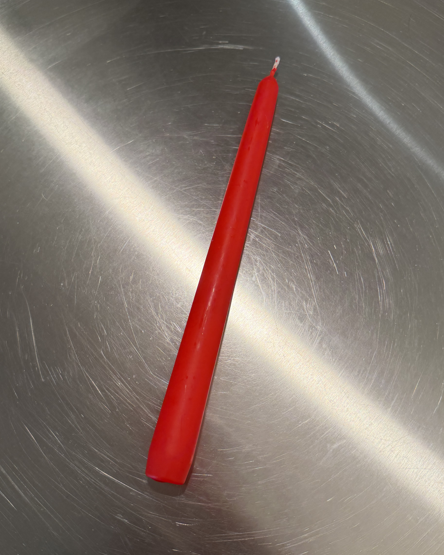 A single red taper candle with a white wick, placed diagonally on a scratched metallic silver surface.