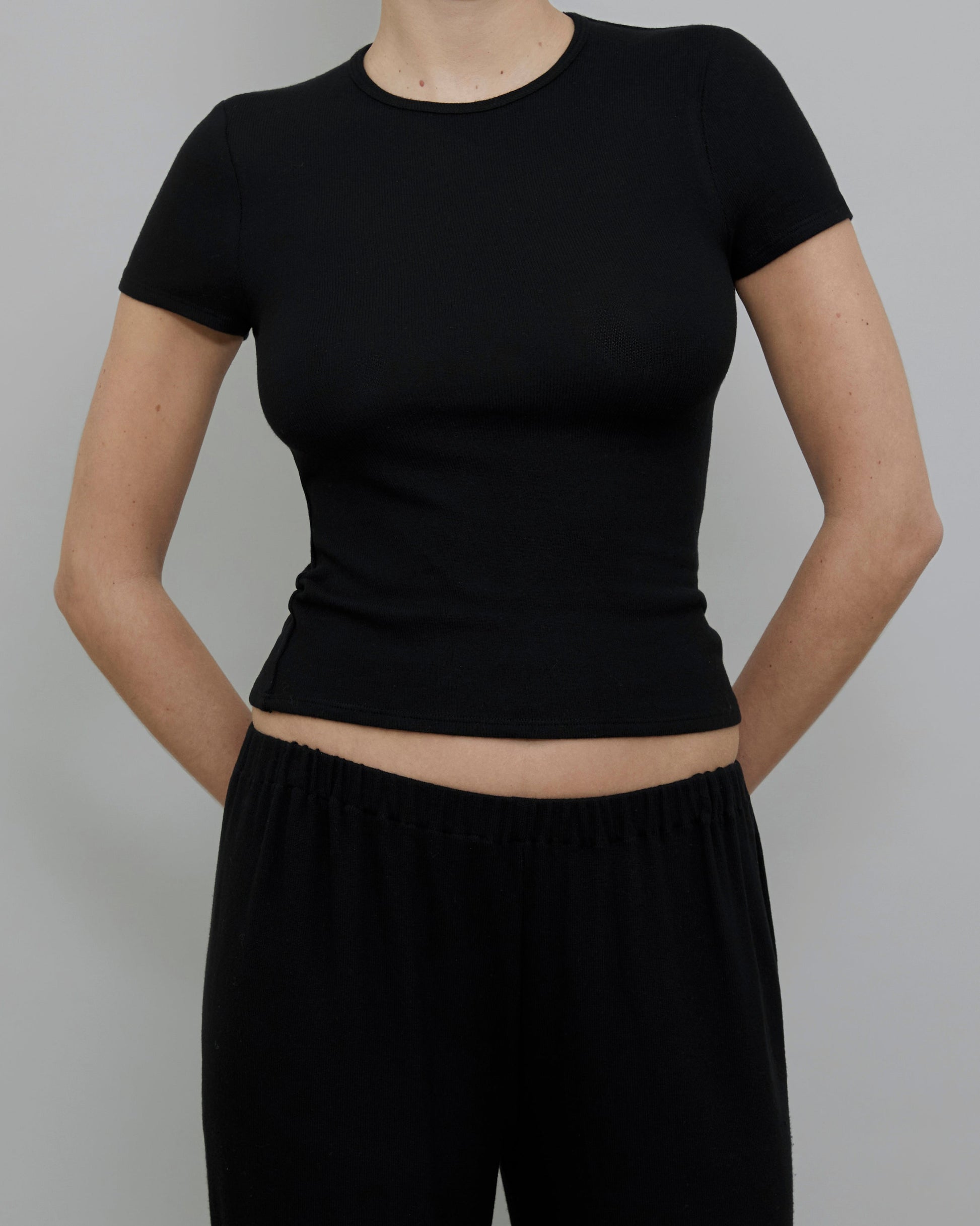 A model wearing a black short-sleeve crewneck crop top and matching black pants, shown from the neck down against a solid gray background.