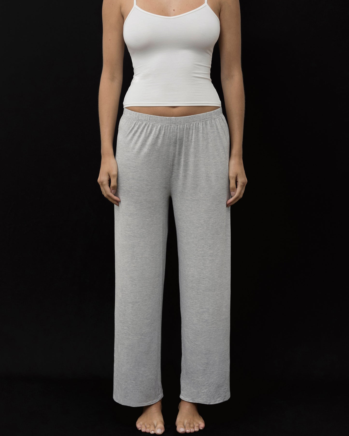 A woman modeling a two-piece loungewear set consisting of a white ribbed camisole top and heather gray wide-leg sweatpants, standing against a solid black background.