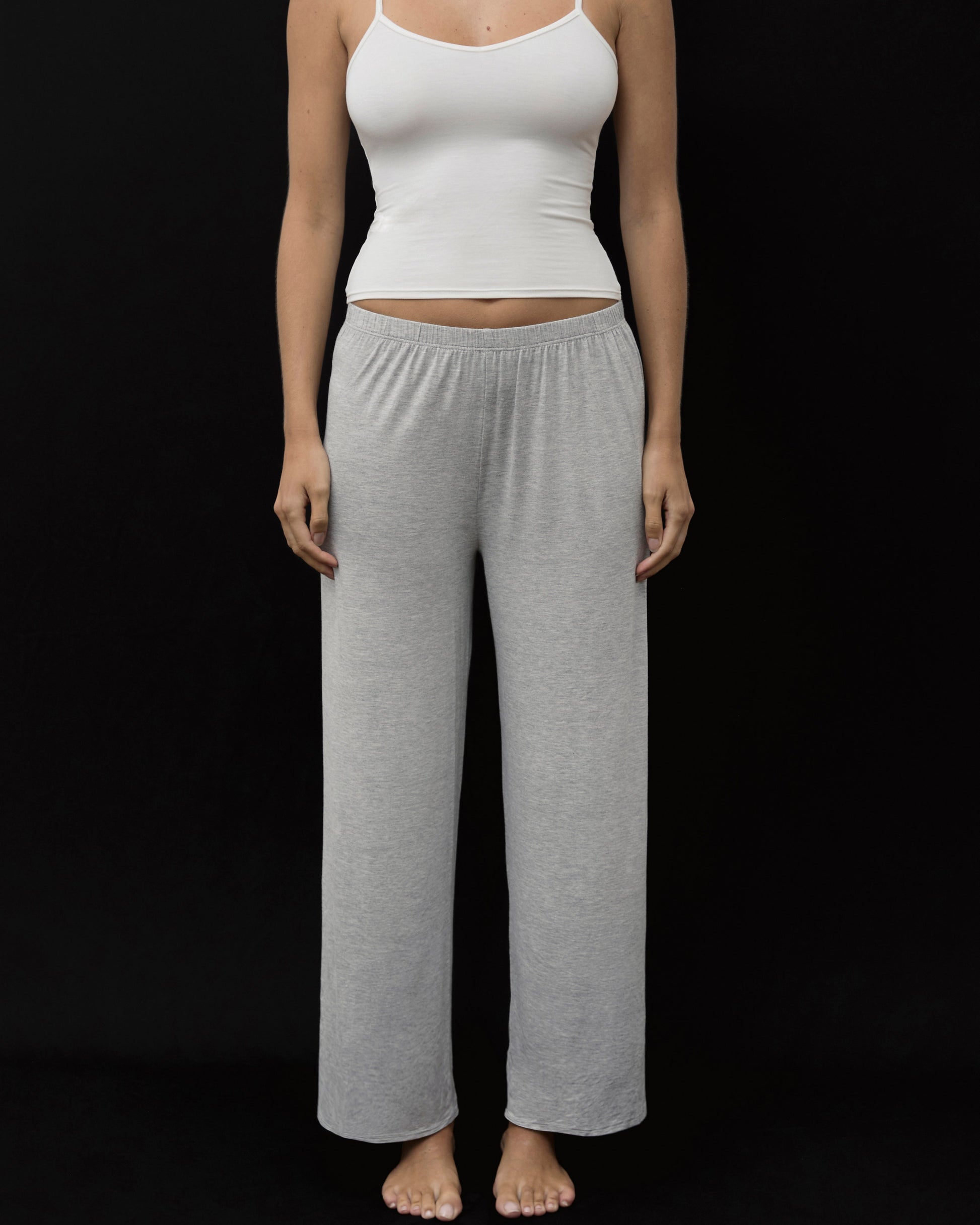 A woman modeling a two-piece loungewear set consisting of a white ribbed camisole top and heather gray wide-leg sweatpants, standing against a solid black background.