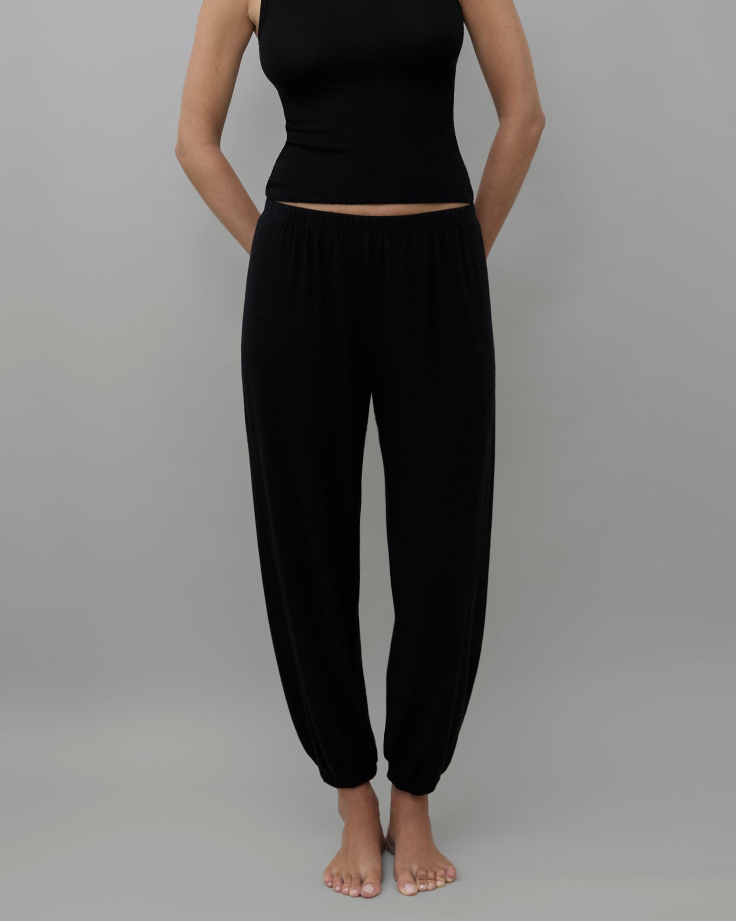 A model wearing a black sleeveless crop top and matching black harem pants, standing against a solid gray background.