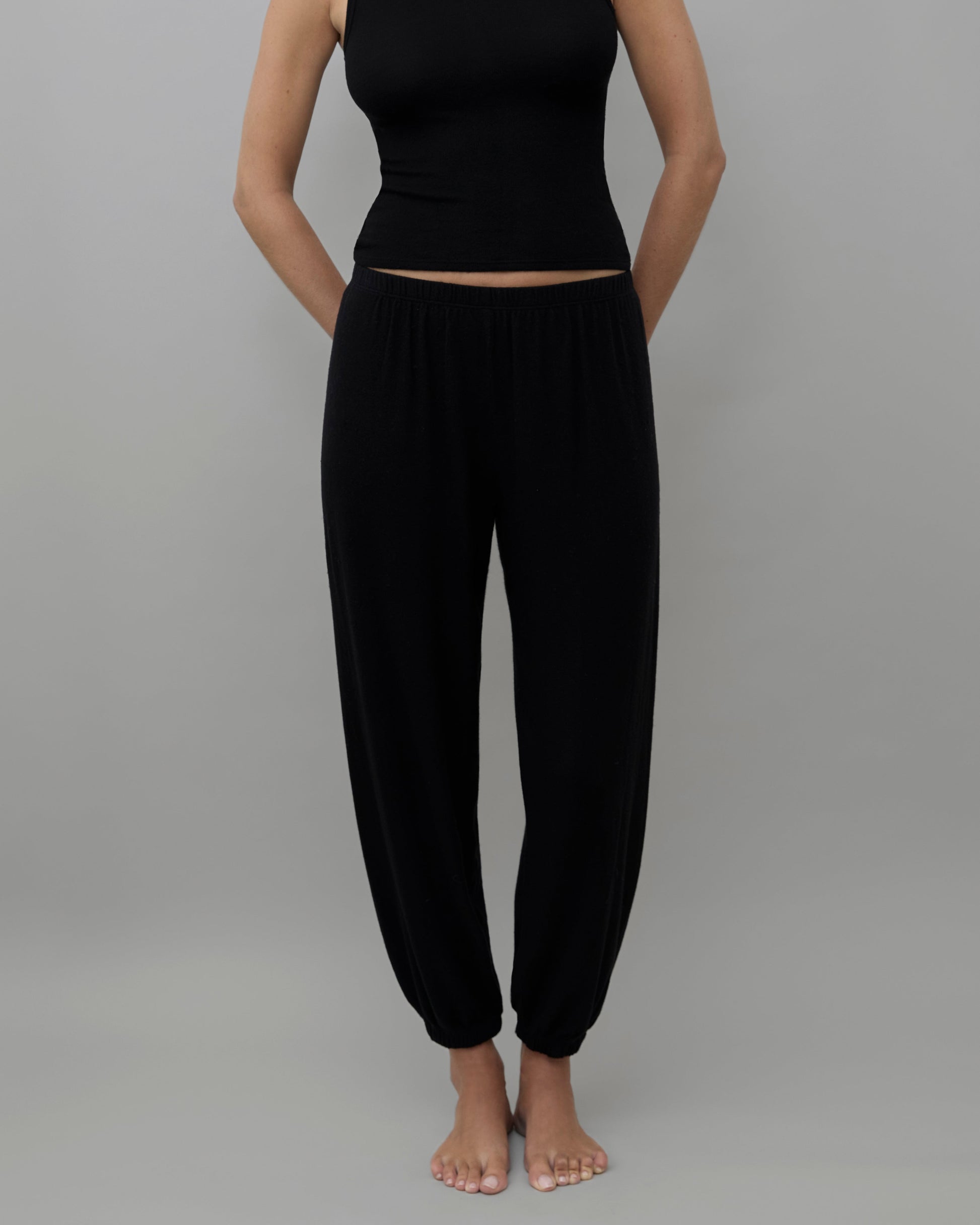 A model wearing a black sleeveless crop top and matching black harem pants, standing against a solid gray background.