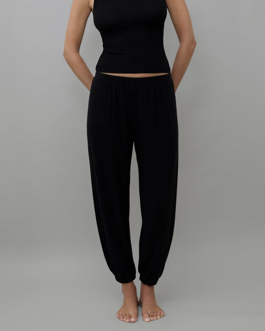 A model wearing a black sleeveless crop top and matching black harem pants, standing against a solid gray background.