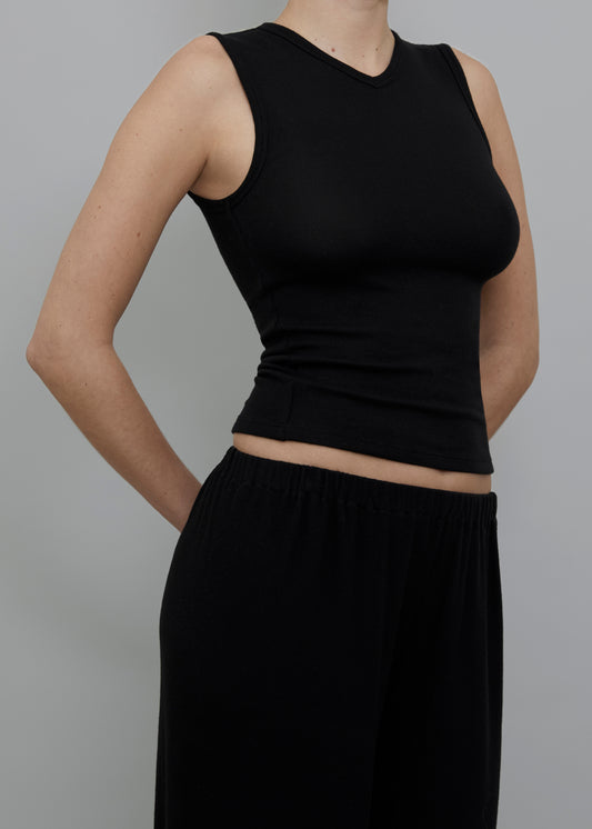 A model wearing a black sleeveless V-neck crop top and matching black wide-leg pants, shown from the neck down against a solid gray background.