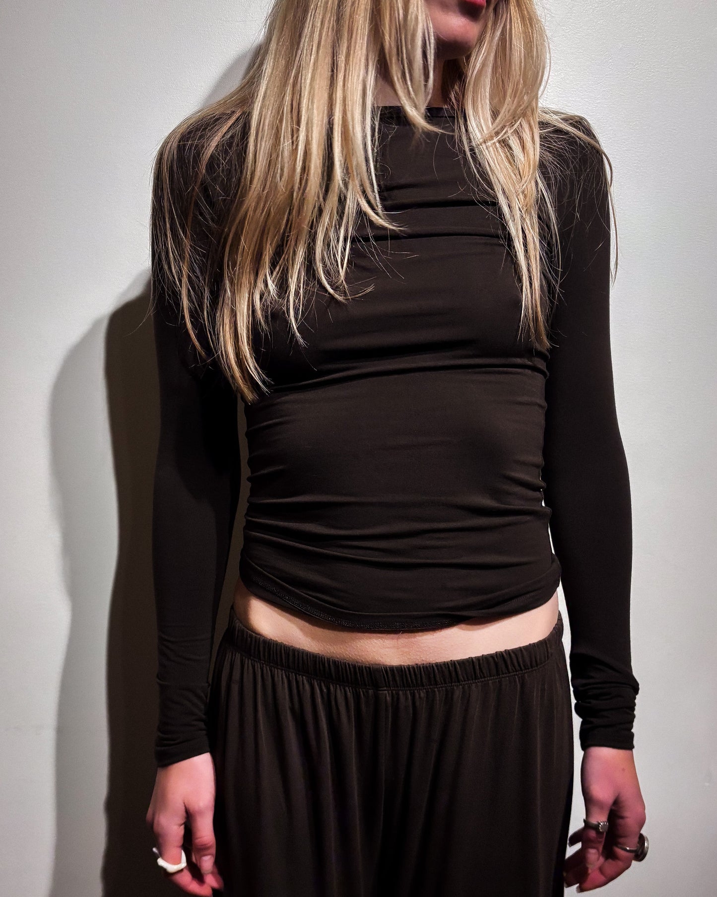 A woman wearing a dark brown long-sleeve ruched crop top and matching dark brown pants, standing against a plain light gray wall.