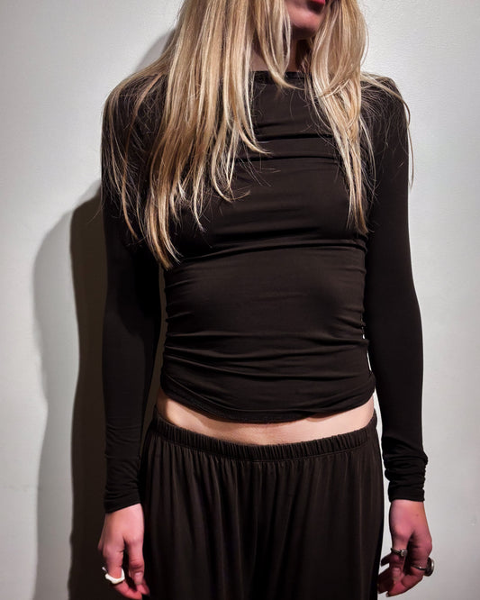 A woman wearing a dark brown long-sleeve ruched crop top and matching dark brown pants, standing against a plain light gray wall.