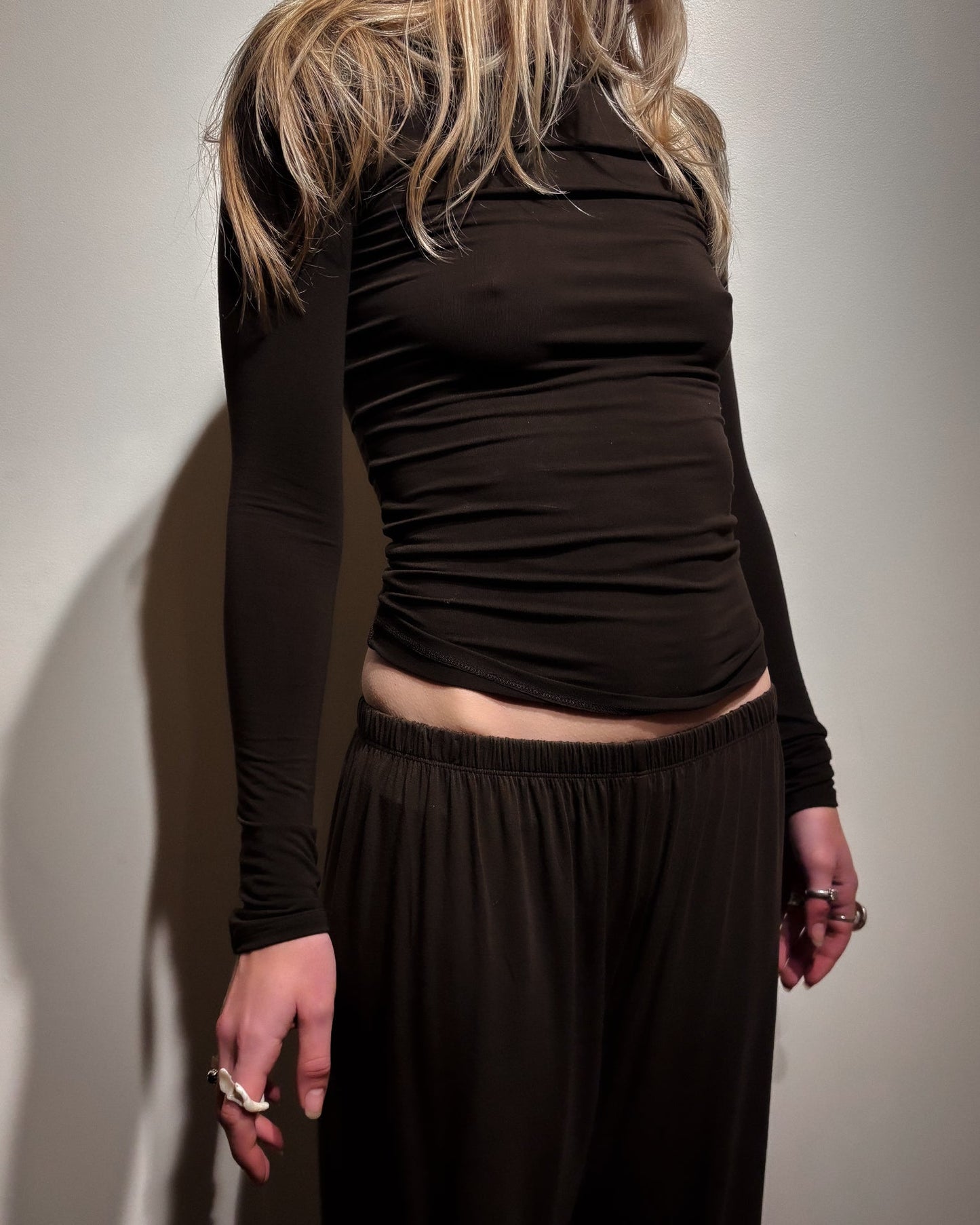 A model wearing a dark brown long-sleeve ruched top and matching dark brown wide-leg pants, shown from a side-back angle against a plain light wall.