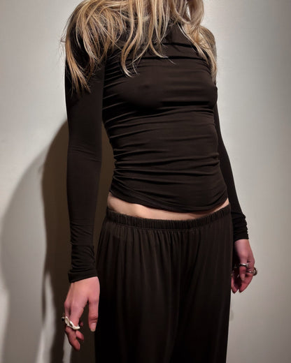 A model wearing a dark brown long-sleeve ruched top and matching dark brown wide-leg pants, shown from a side-back angle against a plain light wall.