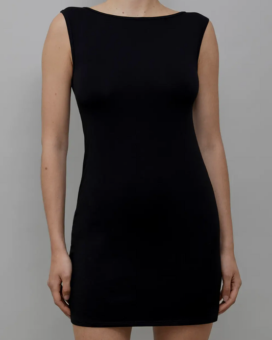 A woman wearing a sleeveless black bodycon mini dress with a boat neckline, shown from the neck down against a plain gray studio background.