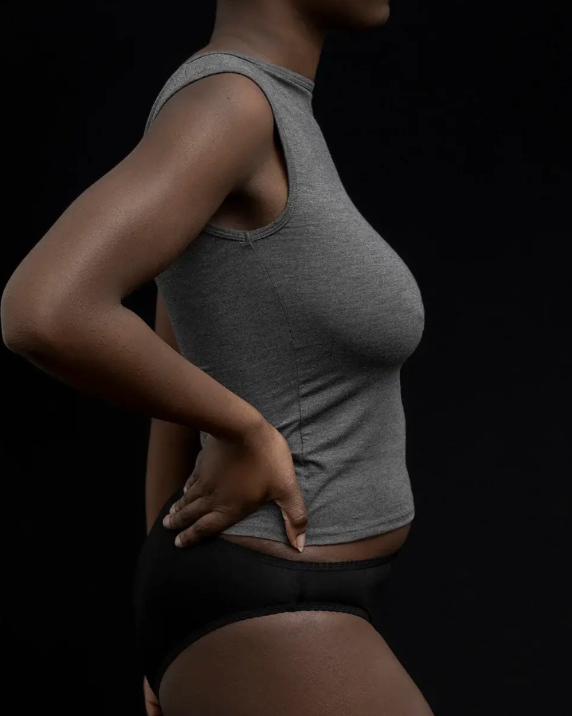 Side profile of a model wearing a heather gray ribbed tank top and black underwear against a solid black background.