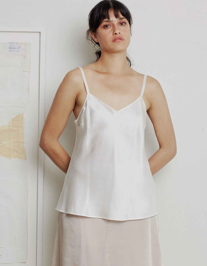 A woman wearing a white silk camisole top with adjustable straps and a gentle v-neckline, paired with a pink skirt.